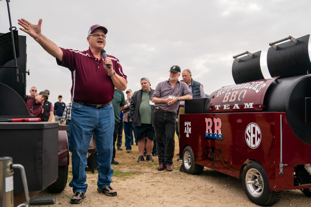 A speaker, Davey Griffin, Ph.D., stands welcoming barbecue enthusiasts who are gathered outside around barbecue pits. 