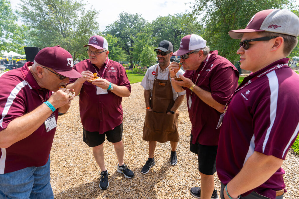 A group of men all dressed in maroon and white trying barbecue. 