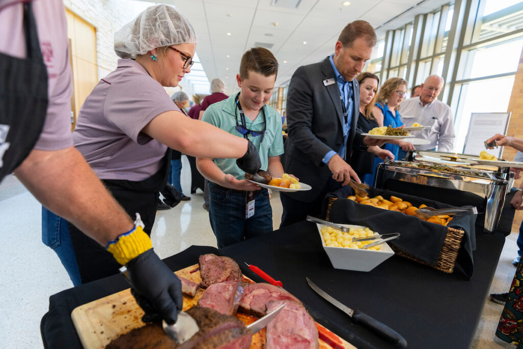 a serving line with people dishing up food and a cutting board full of prime rib at the end