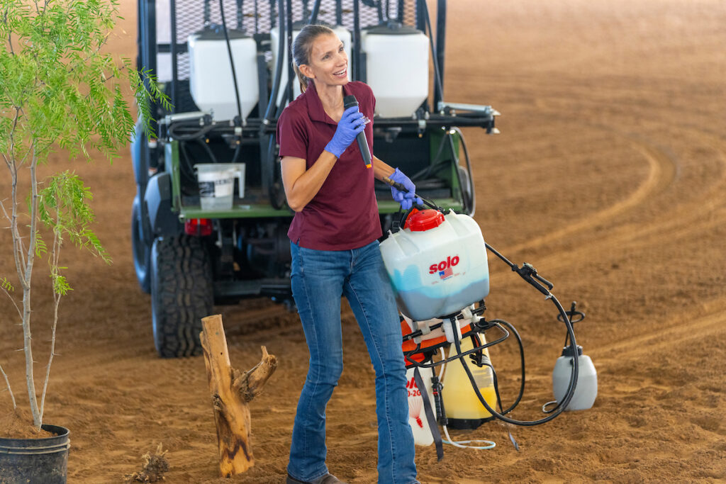 a woman stands on a dirt surface talking into a microphone with a sprayer and canister full of chemicals