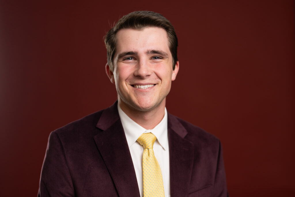 Head and shoulder photo of Zach Hoelscher wearing a maroon coat with a gold tie and a white shirt. There is a maroon background behind him.