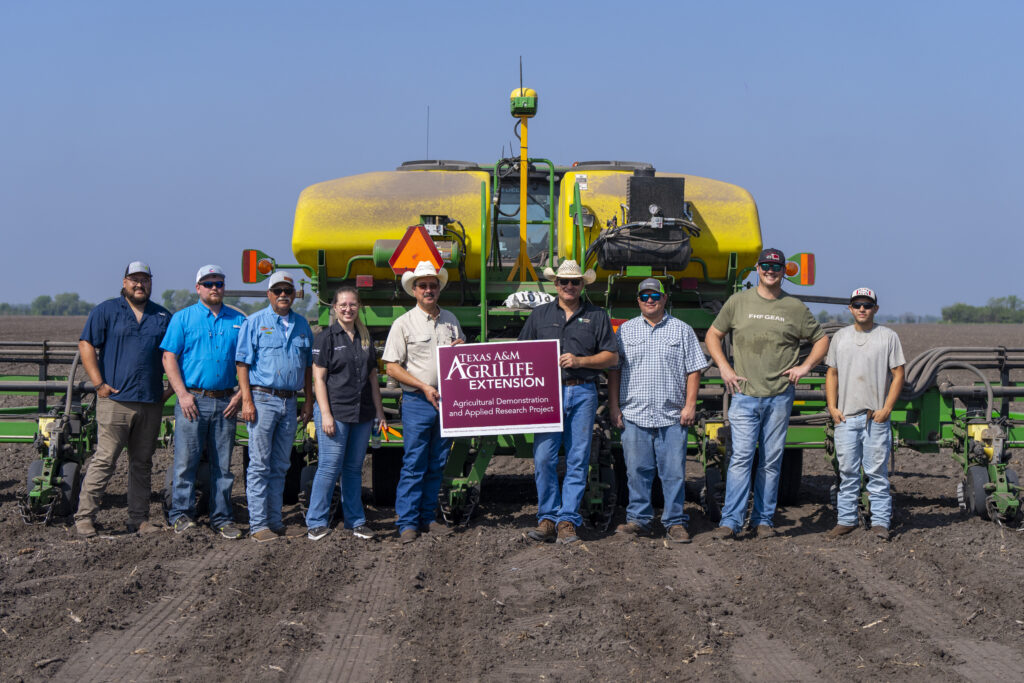 Group photo of farmers and county agents in front of a large piece of farm equipment holding an AgriLife Extension sign 