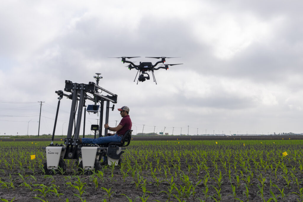 Man in field driving latest version of farm equiment with drone flying overhead 