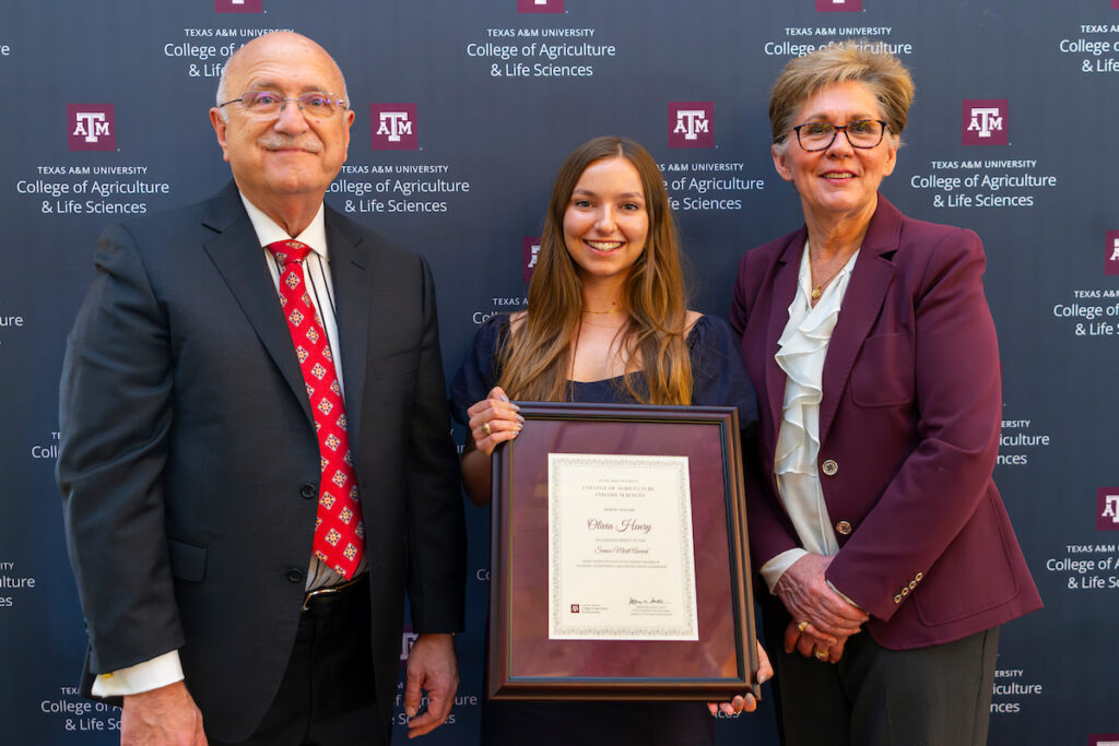 A young woman stands between an older man and older woman with a Texas A&M backdrop behind them. She is holding a plaque she was awarded for leadership.
