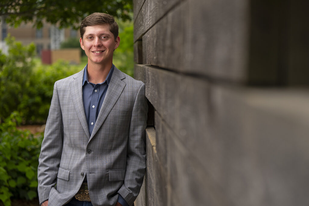 Kaden Simon '26, recipient of the Hullabaloo-U Peer Mentor of the Year award, leans against a brick wall while wearing a grey suit and blue button-down shirt.