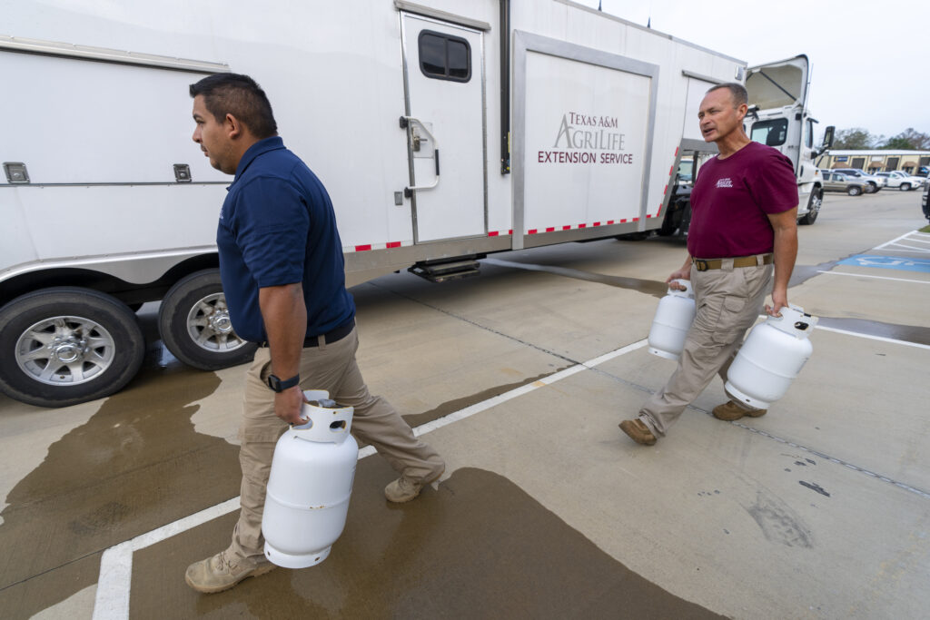 Two Disaster agents carrying propane tanks prepare to load them into a white trailer with the words Texas A&M AgriLife Extension Service on it. Both men are wearing khaki pants and one has a blue short and the other is wearing a maroon shirt. 