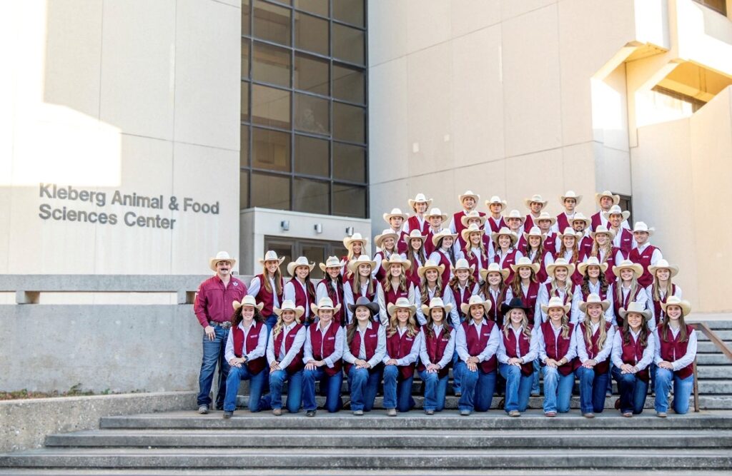Five rows of students either kneeling or standing on the steps of the Kleberg Animal & Food Sciences Center all dressed in jeans and maroon vests over blue long-sleeved shirts, with one coach in a maroon shirt.