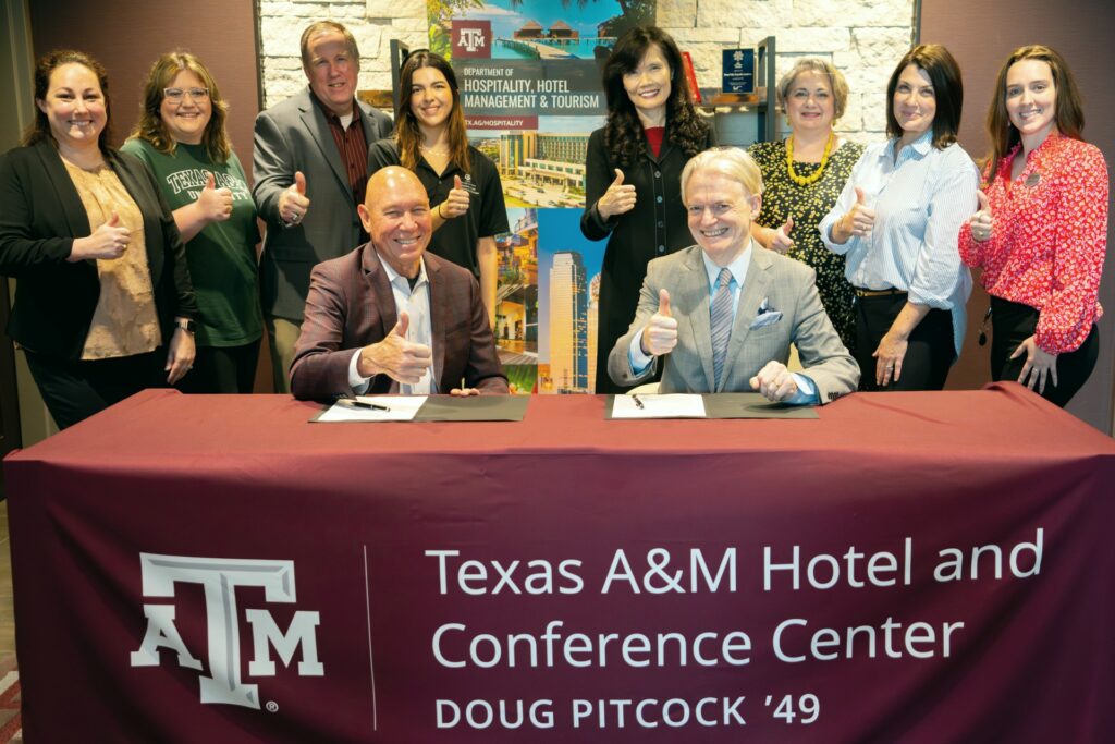 Ten individuals, seven women and three men, giving a thumbs up. Two of the men are sitting at a table with a tablecloth that have the ATM logo and the words Texas A&M Hotel and Conference Center Doug Pitcock '49. The other eight individuals are standing behind them.
