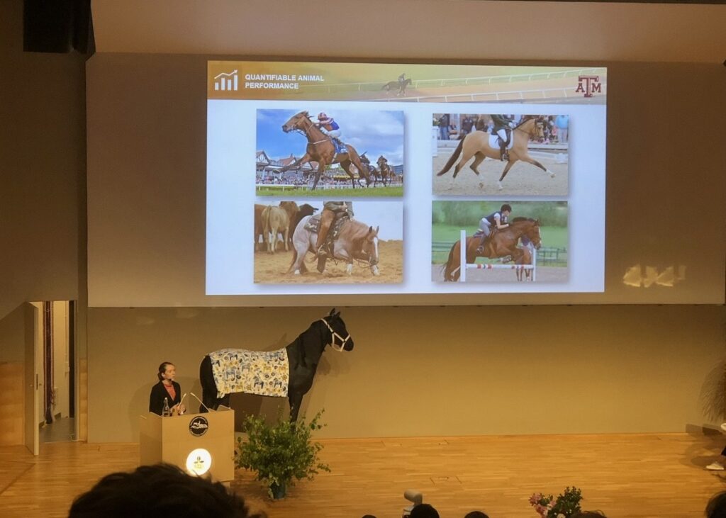 A woman, Lauren Wesolowski, standing behind a podium in the corner with a life-size statue of a horse behind her and a large screen above her showing four pictures of horse activities
