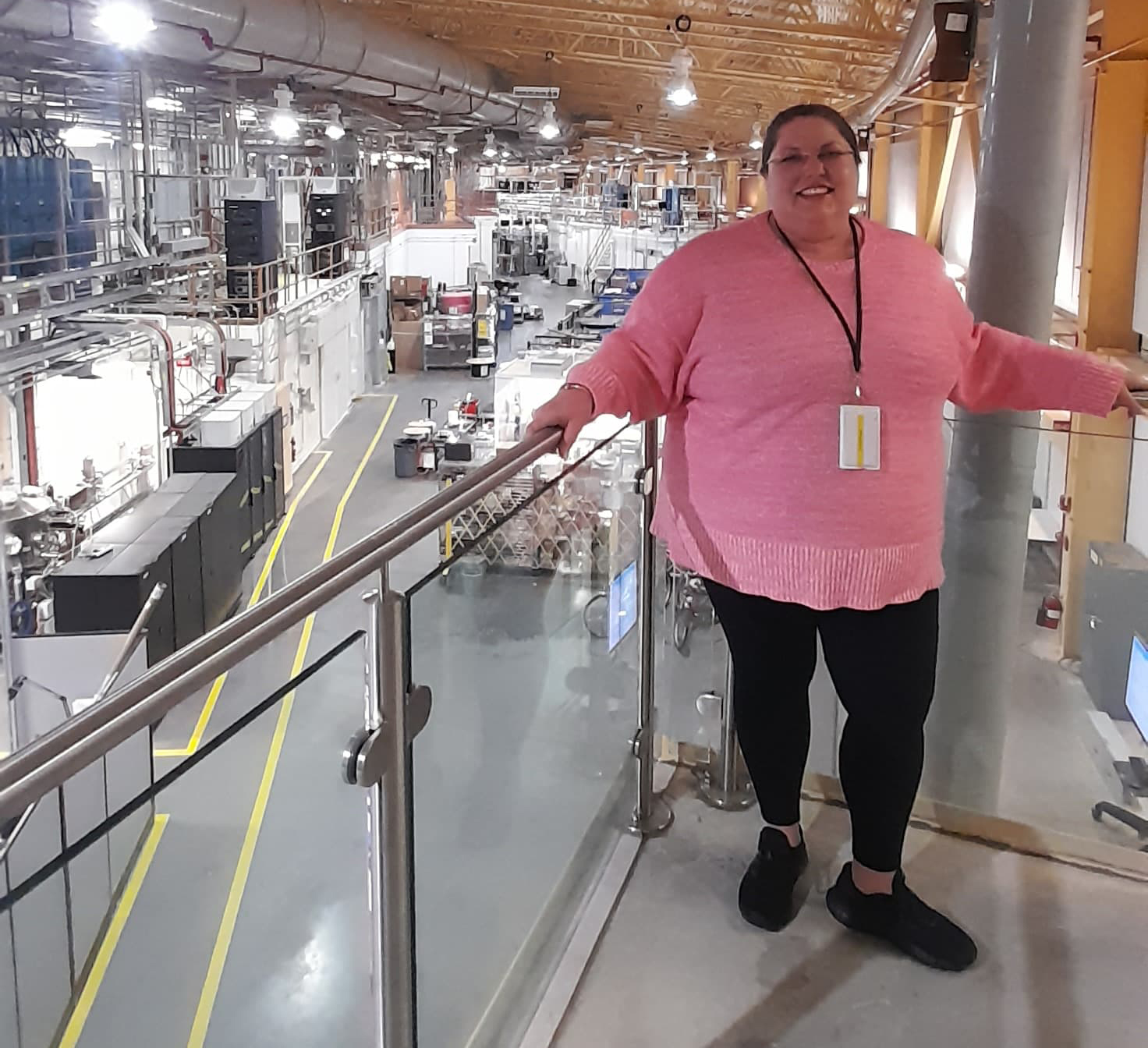 A woman in a pink sweater stands on a walkway above a large laboratory that stretches far behind her with lots of equipment