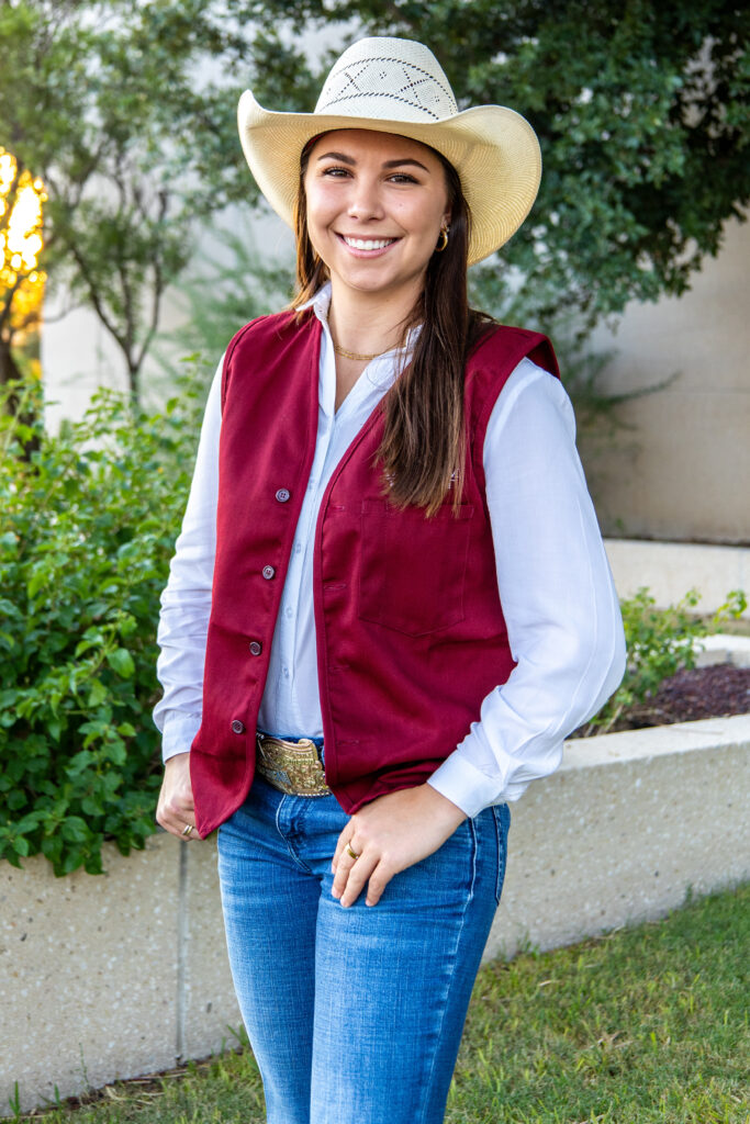 A smiling woman standing outdoors in blue jeans and a maroon vest with a straw cowboy hat on 