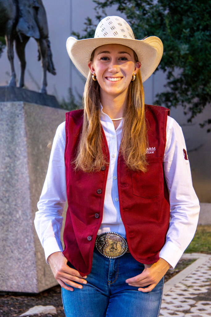 A woman standing outdoors in blue jeans and a maroon vest with a straw cowboy hat on and a big belt buckle showing