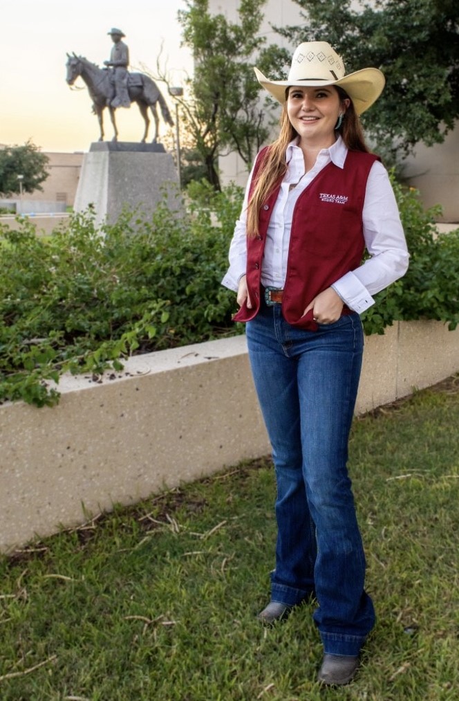 A woman standing outdoors in blue jeans and a maroon vest with a straw cowboy hat on