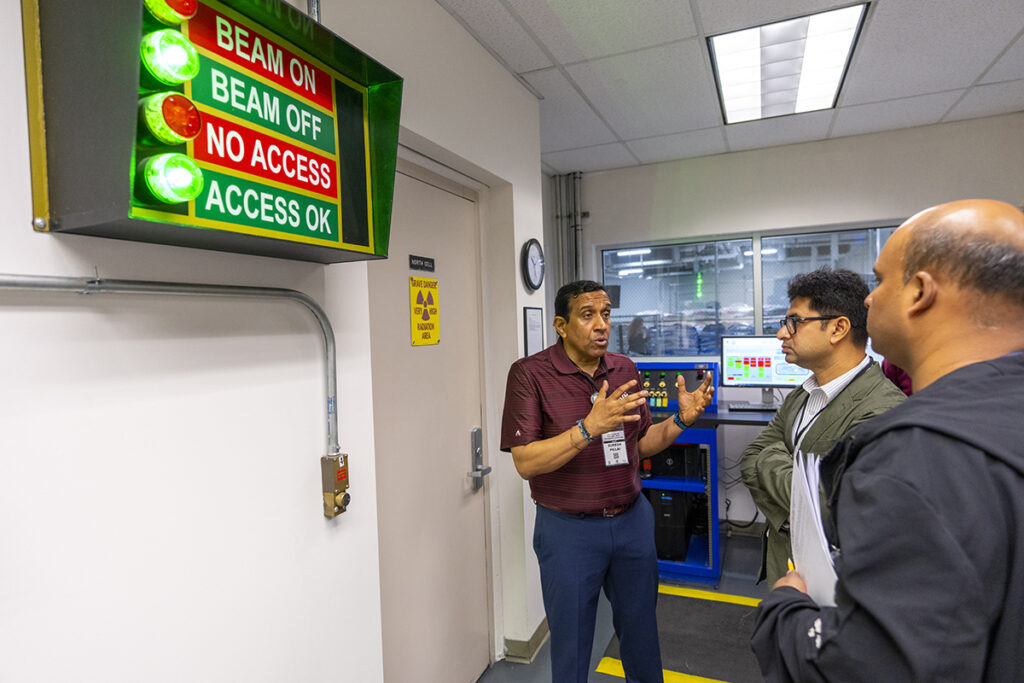 Three men stand in front of a door and a sign with red and green lights read: Beam On, Beam Off, No Access and Access OK.