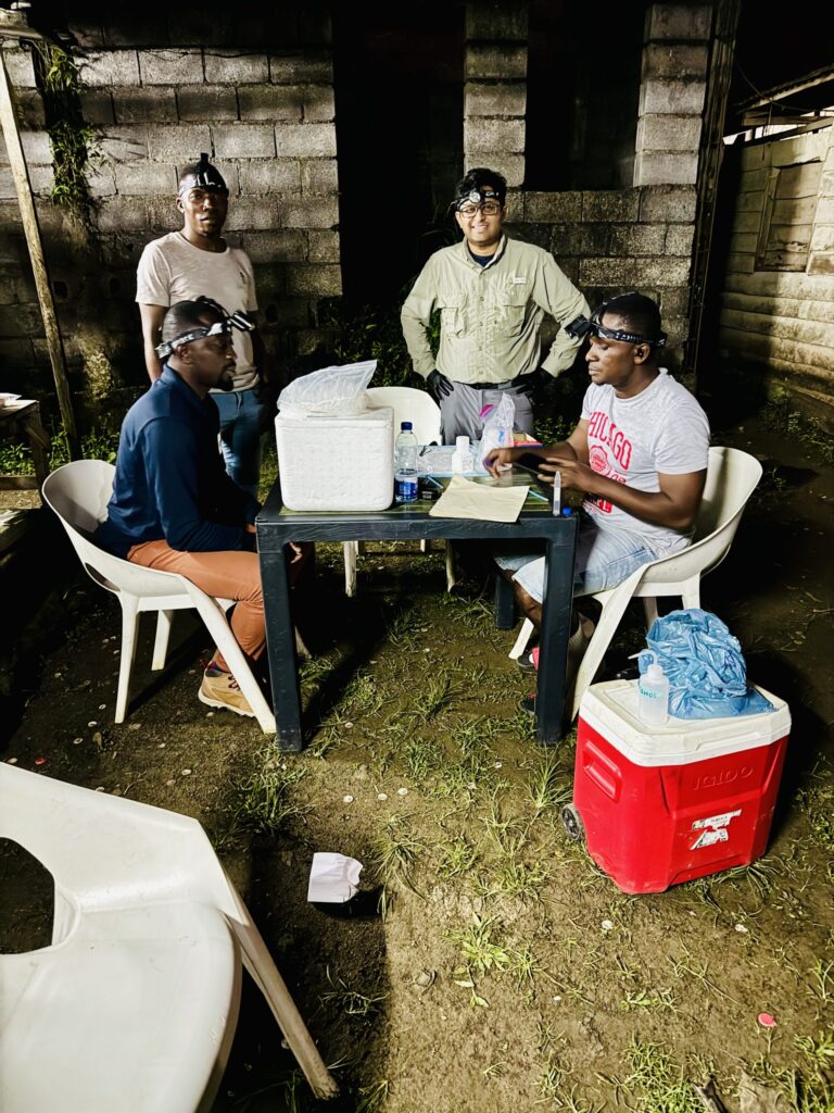 Giri Athrey, Ph.D., stands with several other men who are part of the Equatorial Guinea team that is working to eradicate malaria. They stand outdoors at night around a table working on samples to be sent to a lab.