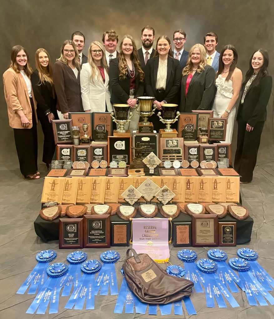 A group of students on the Meat Judging team standing together with a row of awards layed out on the floor in front of them.