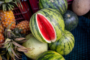 A cut open green and red watermelon sitting on top of other water melons and surrounded by other fruits all laying on a table.