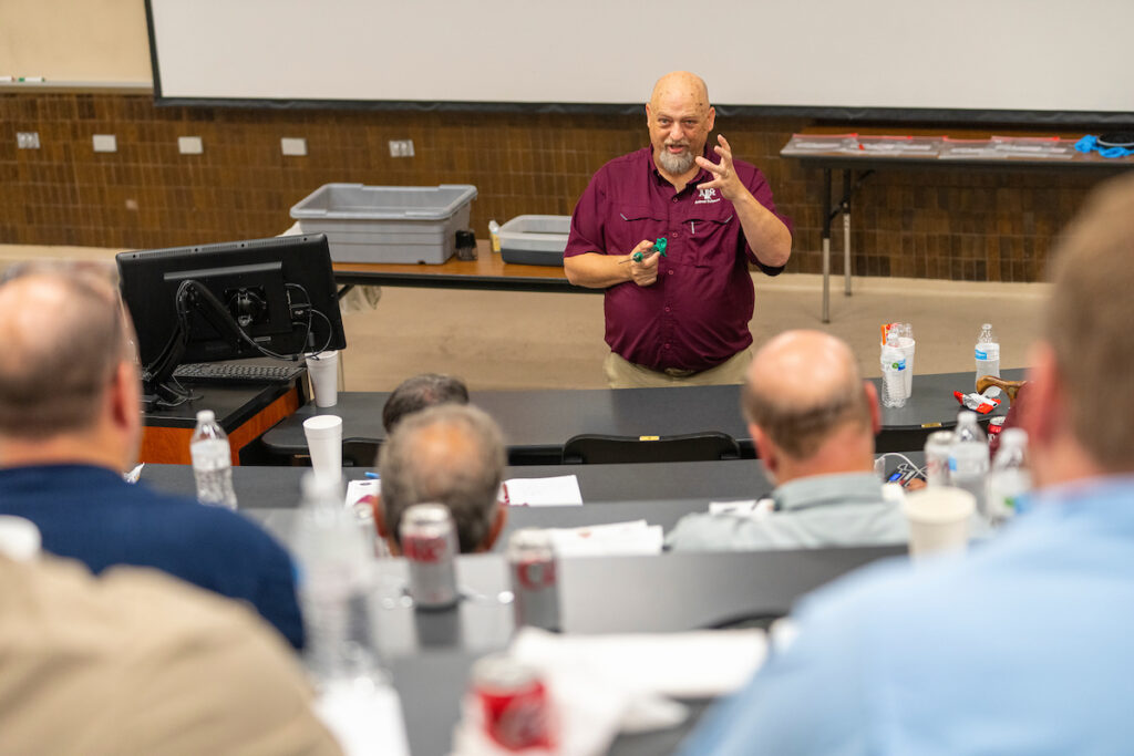 A man in a maroon shirt stands at the front of a classroom talking with a marinade tube.