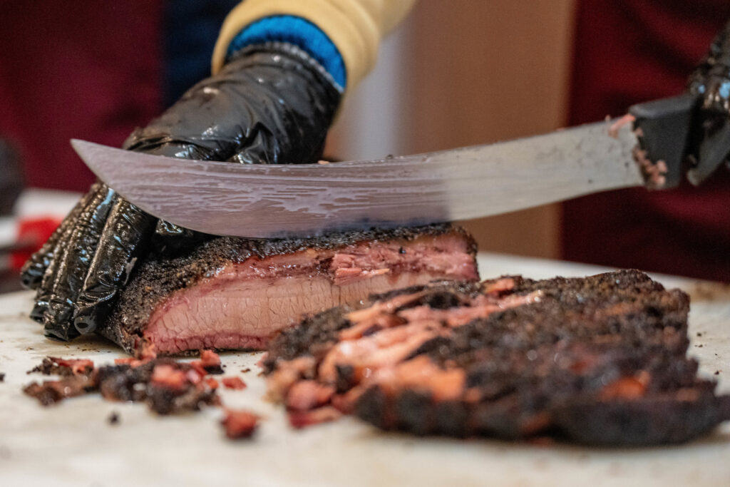 One person holding a cut of brisket meat, cutting into it with a long knife and gloves on.