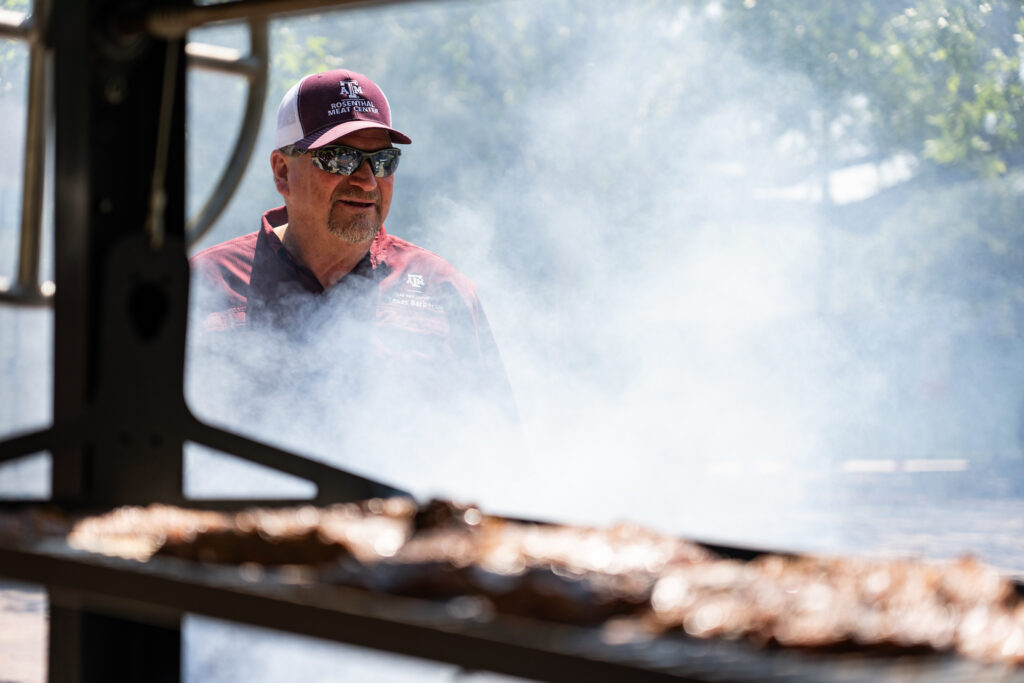 Ray Riley, standing behind a grill with smoke surrounding him and grilling steaks on the grill in front of him.