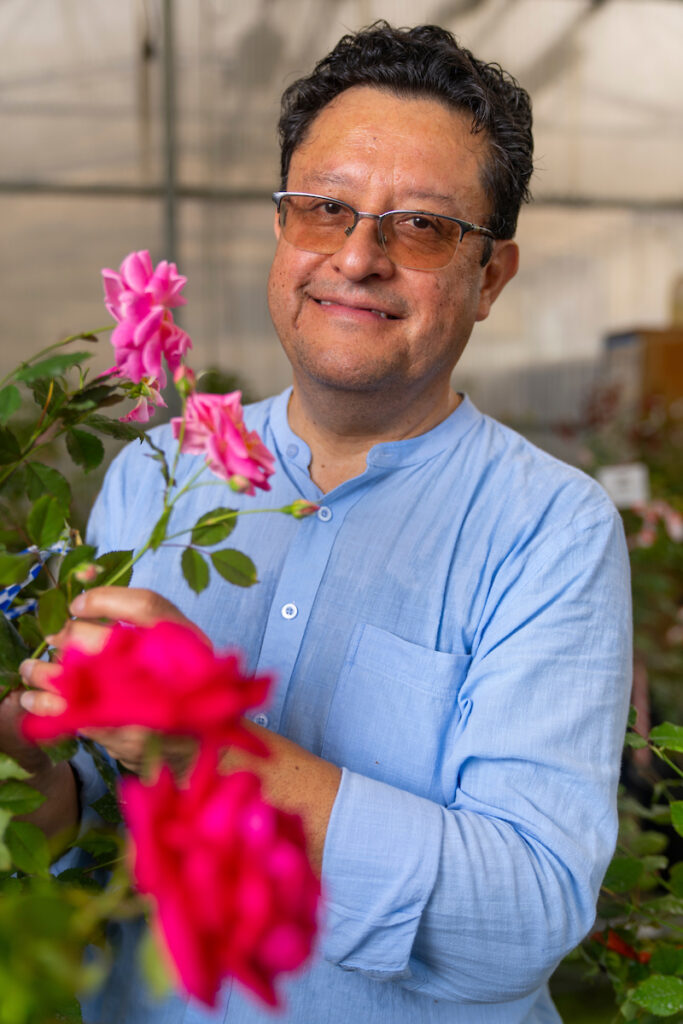 A man, Oscar Riera-Lizarazu, Ph.D., stands holding a pink rose bush. he is wearing a blue long sleeve shirt