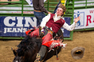 A competitor in a maroon Texas A&M vest and red chaps leans back as he competes in bareback riding on a black horse