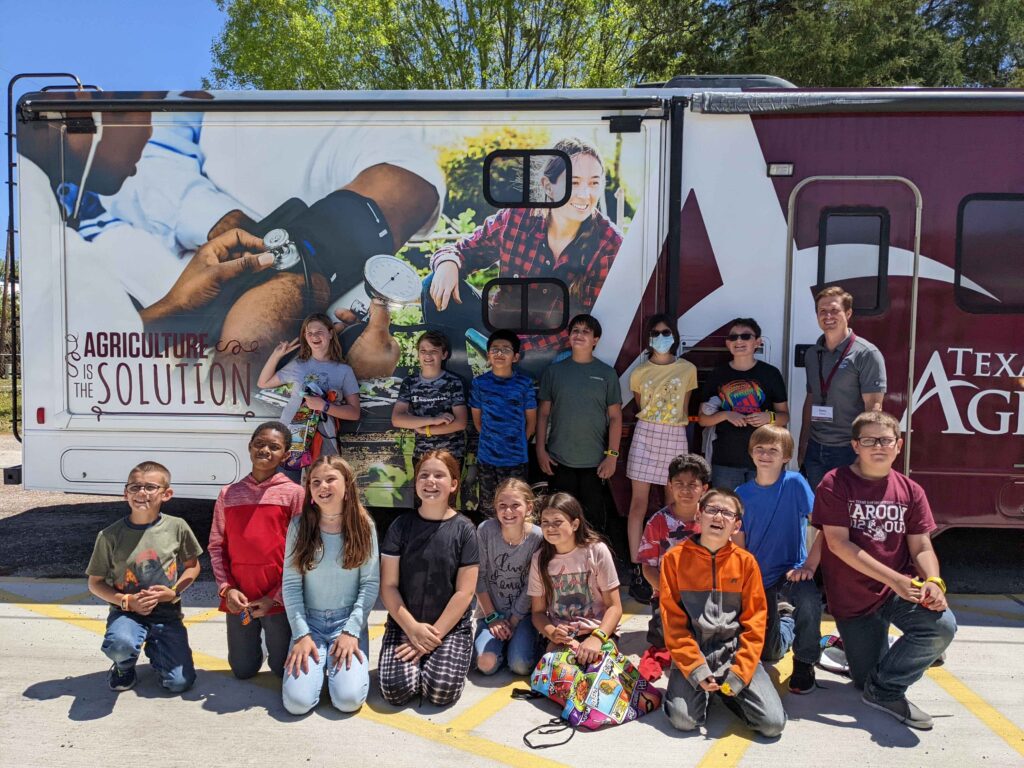 A group of smiling kids poses in front of a mobile health assessment vehicle with a Texas A&M AgriLife staff memeber.