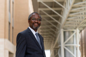 Dr. Henry Fadamiro stands smiling in a navy suit and blue toe in an outdoor corridor at Texas A&M.