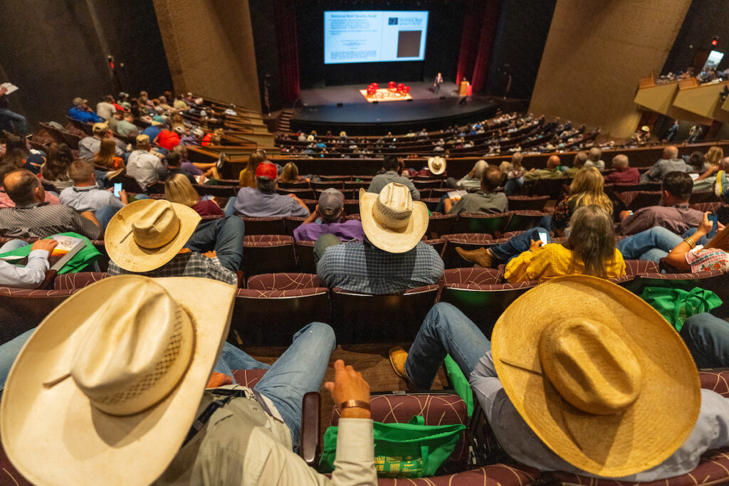 A large Texas A&M auditorium filled with men and women, many in cowboy hats, listening to a speaker on stage with a beef cattle presentation during the Beef Cattle Short Course.