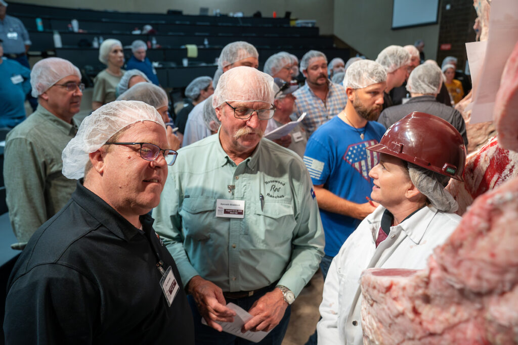 a bunch of men with hair nets on and a woman with a hard hat on stand in a classroom setting with hanging beef carcasses as a part of Beef 706