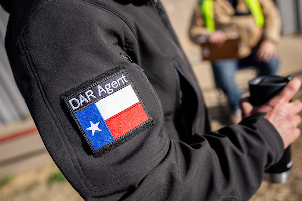 The right arm of an individual wearing a jacket with a Disaster Assessment and Recovery badge on it. The individual is also holding a cup in their right hand. 