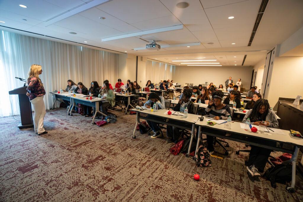 A woman stands at the front of a room speaking to students that are sitting at rectangular tables.