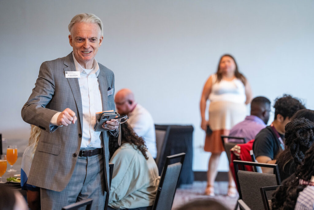 Brian King, Ph.D., head of the Department Hospitality, Hotel Management and Tourism, stands in a grey suit in the middle of a room of students sitting at round tables.