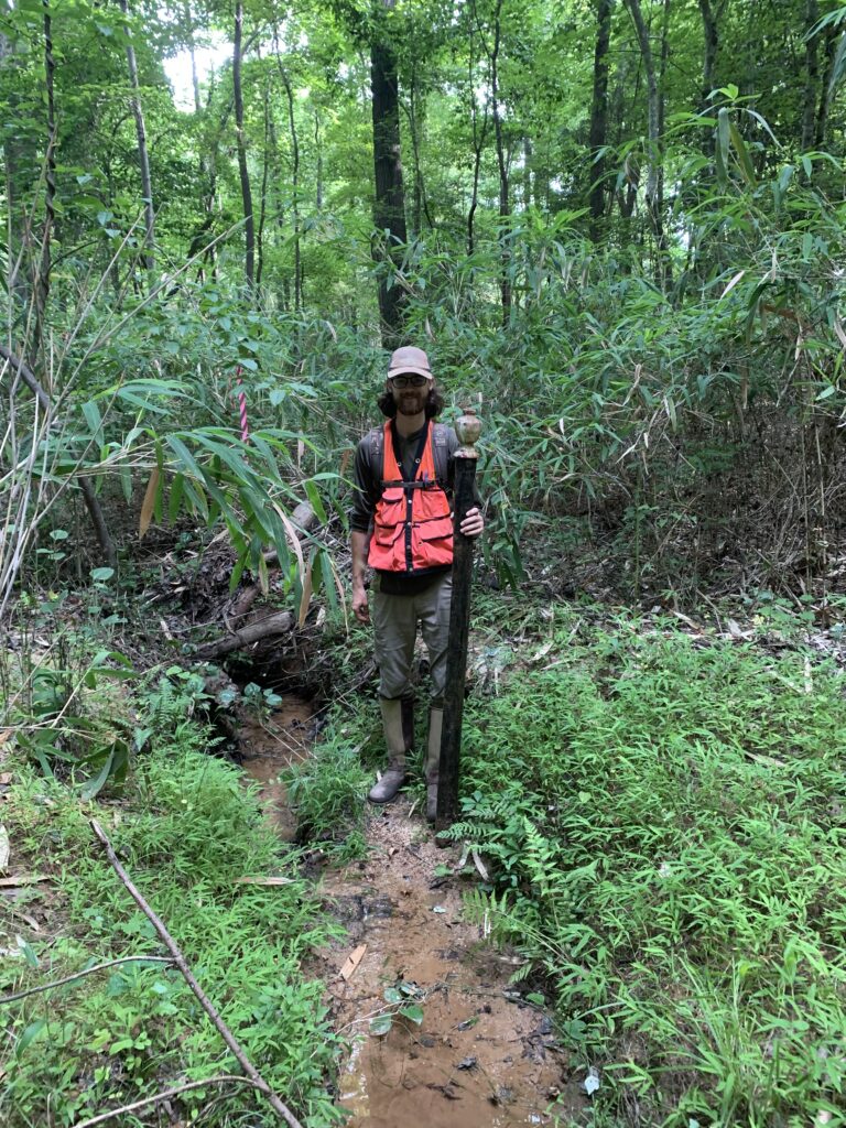 A man wearing an orange vest stands with a large pole in the forest. 