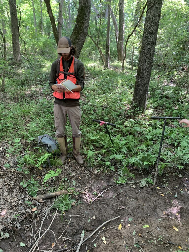 A man wearing an orange vest stands while writing in a notebook in a forest.