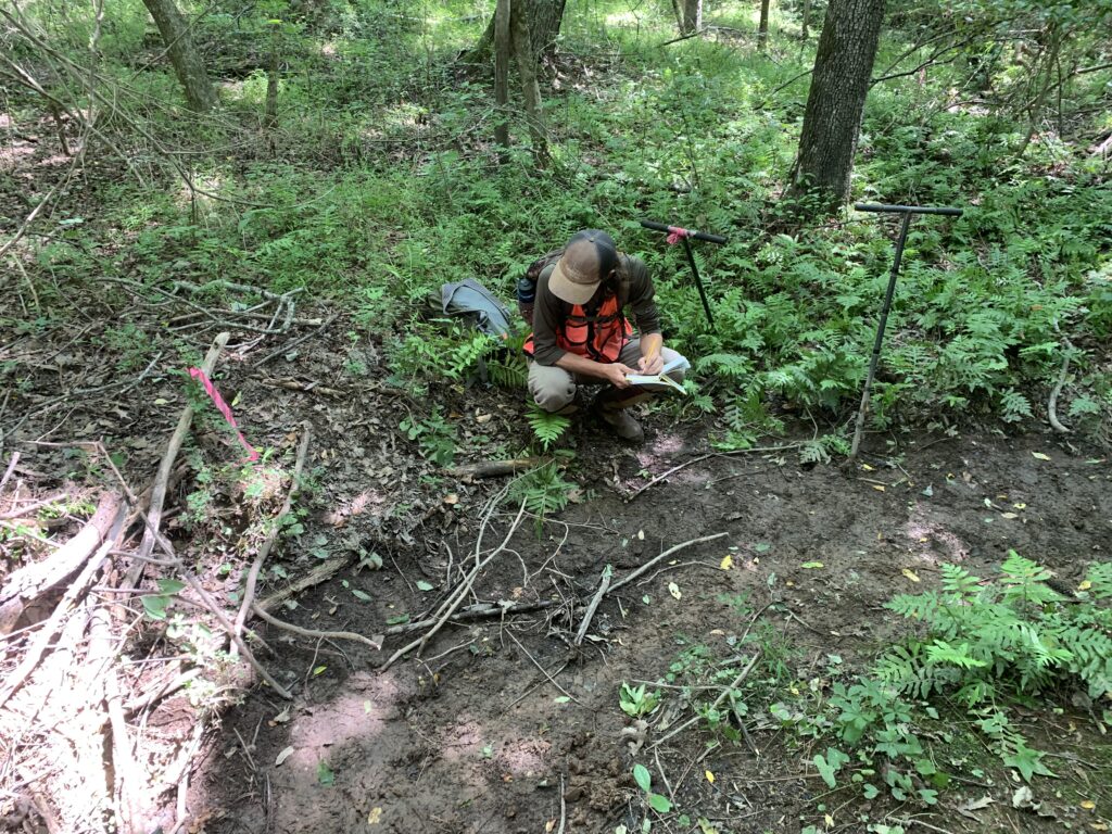 A man wearing an orange vest squats with while writing in a notebook in the forest. 