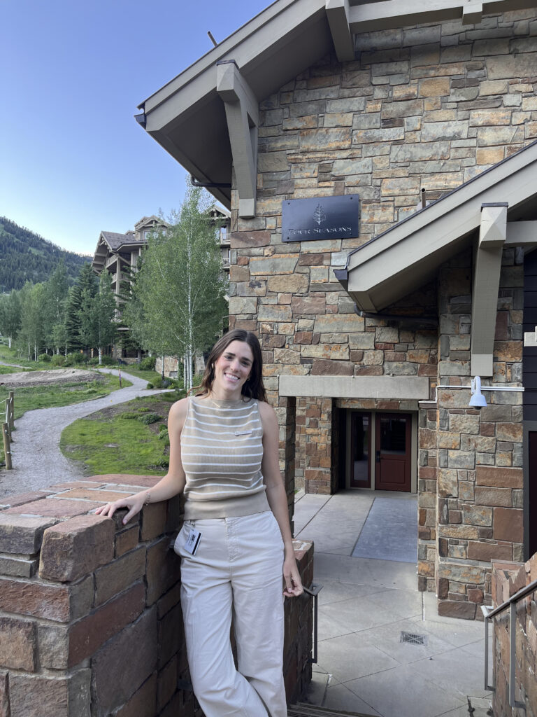 A woman wearing a striped tan and white tank top and tan pants stands in front of trees and a stone building. 