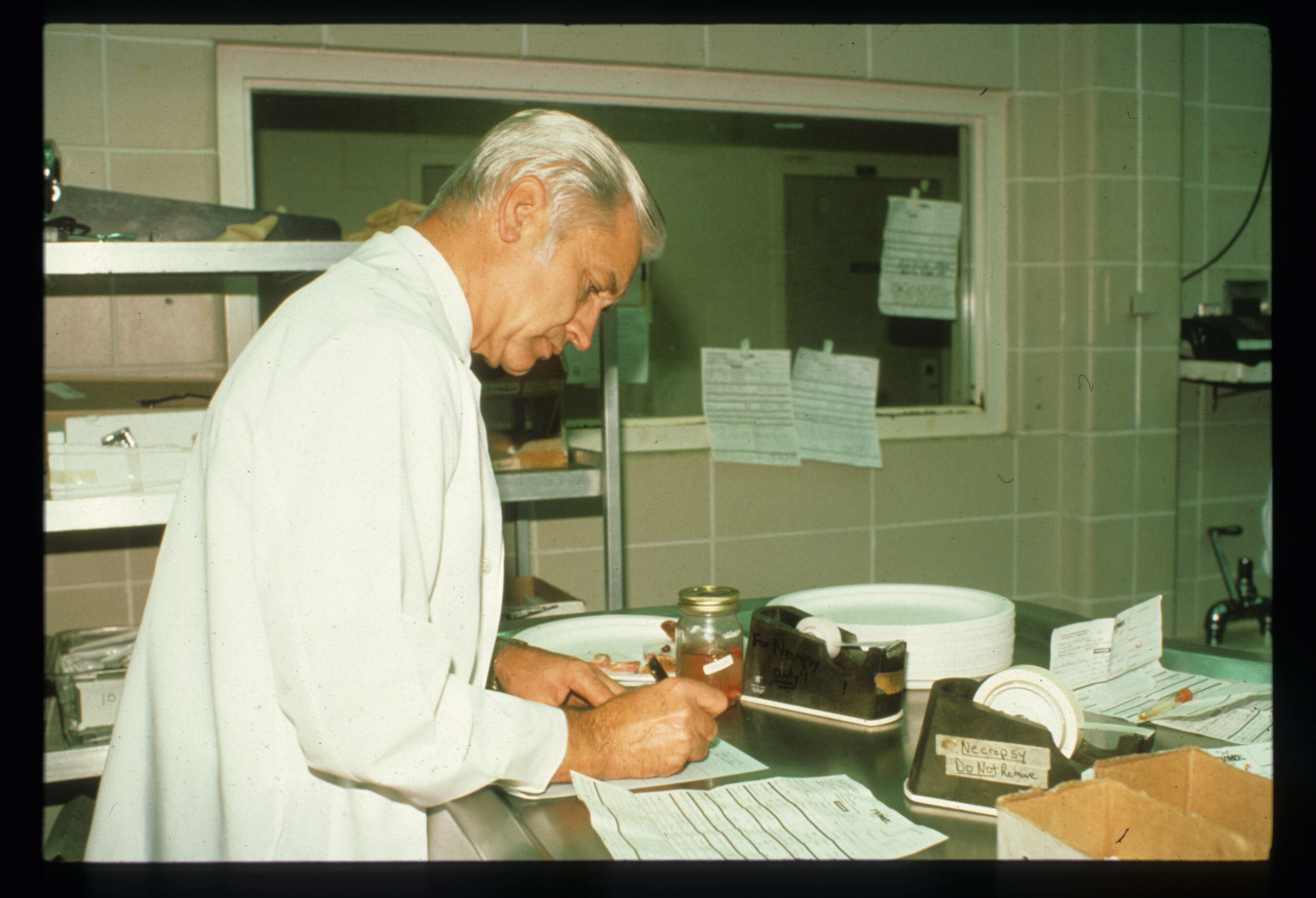 A man in a lab coat at a lab table, writing on papers in front of him. There are various items on the table. 