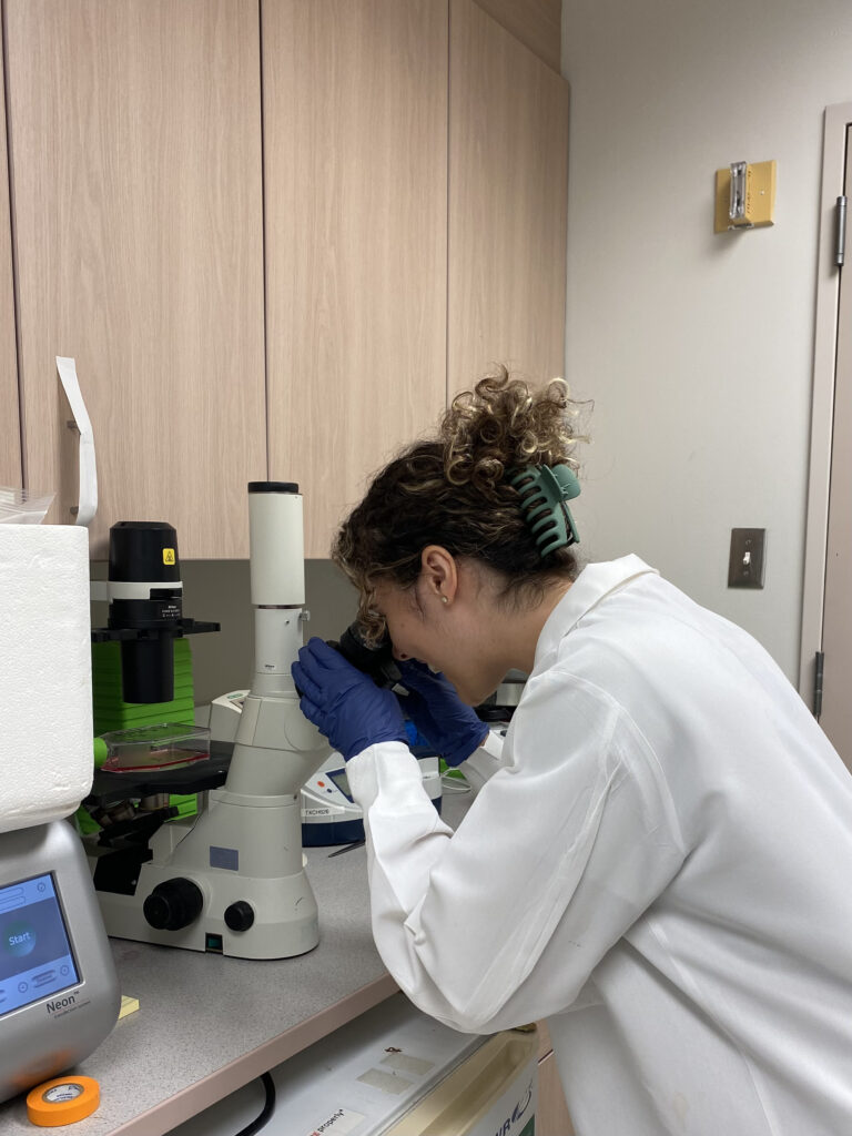 A woman in a white lab coat looks through a microscope. 