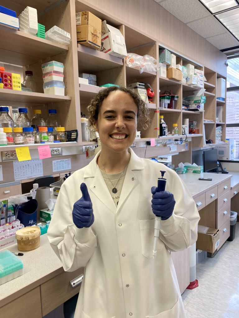 A woman in a white coat and blue latex gloves stands with both thumbs up in a lab. 