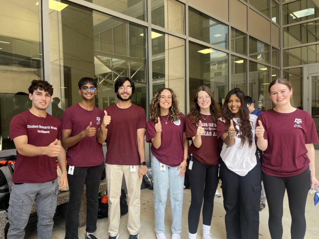 Three men and two women stand  in a line with their thumbs up. 