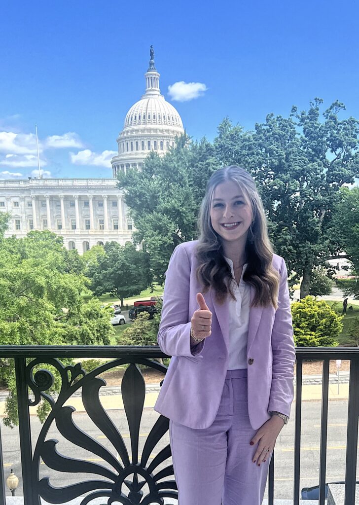 Zoë Meeks, who is completing an internship at the National Cotton Council, stands in a purple suit while giving a thumbs up movement in front of the Washington, D.C., capitol building. 