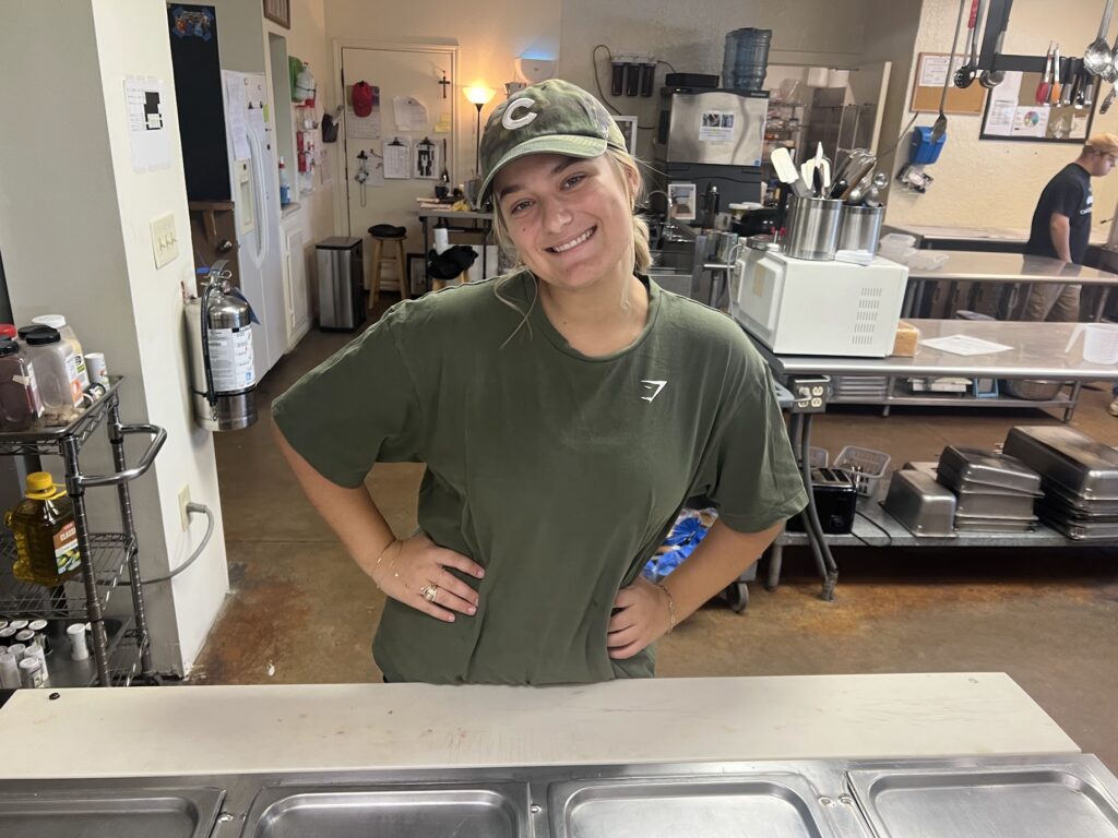 A woman wearing a green t-shirt and a green hat stands with her hands on her hips in a kitchen. 