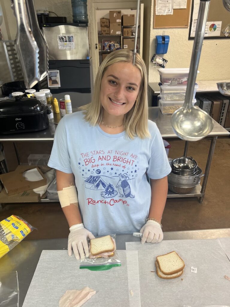 A woman wearing a light blue t-shirt and white latex gloves stands in a kitchen while making sandwiches. 