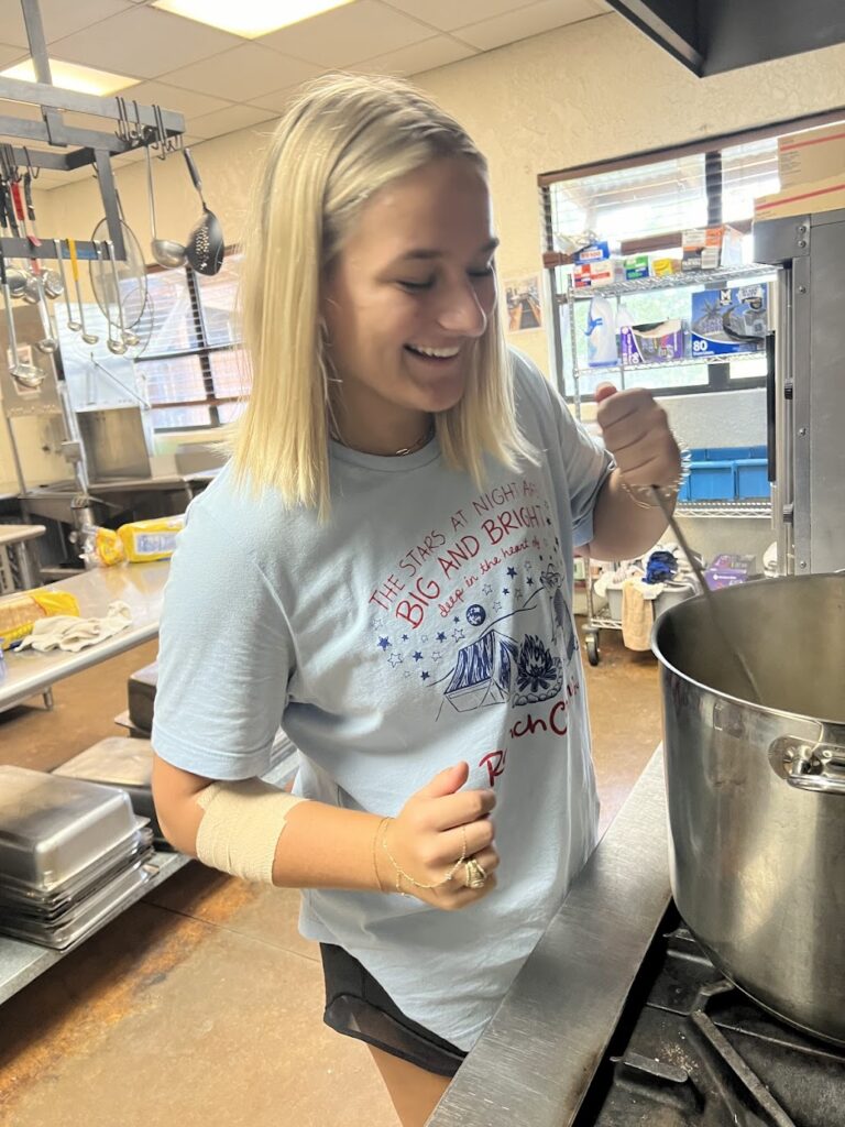 A woman wearing a light blue t-shirt stirs a ladle in a large metal pot in a kitchen.