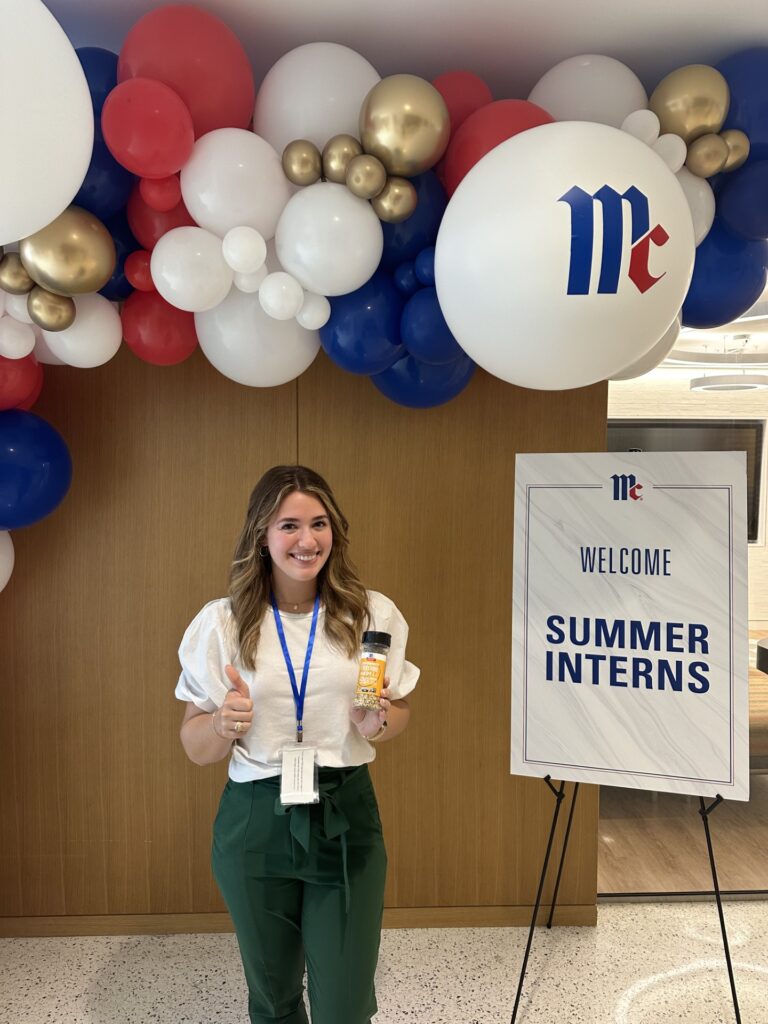 A woman wearing a white shirt and green pants stands with her thumb up next to a welcome sign for McCormick summer interns. 