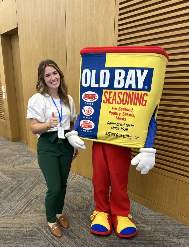 A woman wearing a white shirt and green pants stands with her thumb up next to an old bay seasoning mascot. 