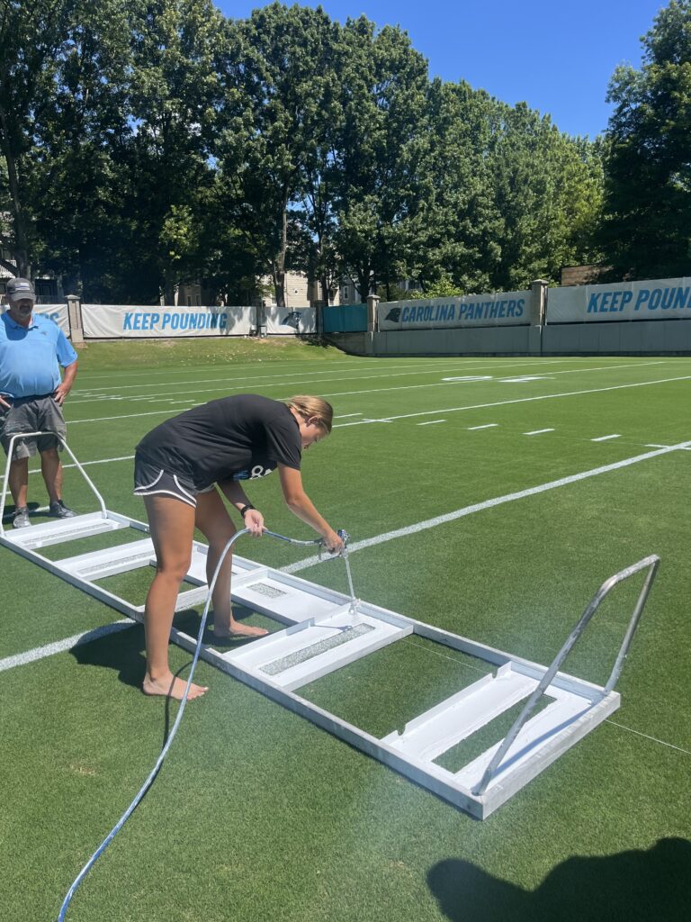 A woman in a black shirt and black short uses large stencils to paint white lines on a football field