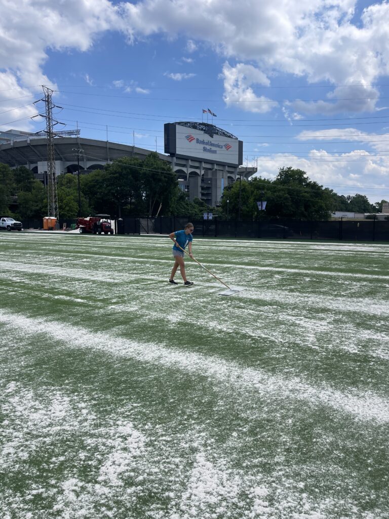 A woman uses a rake to apply white fertilizer to a football field. 