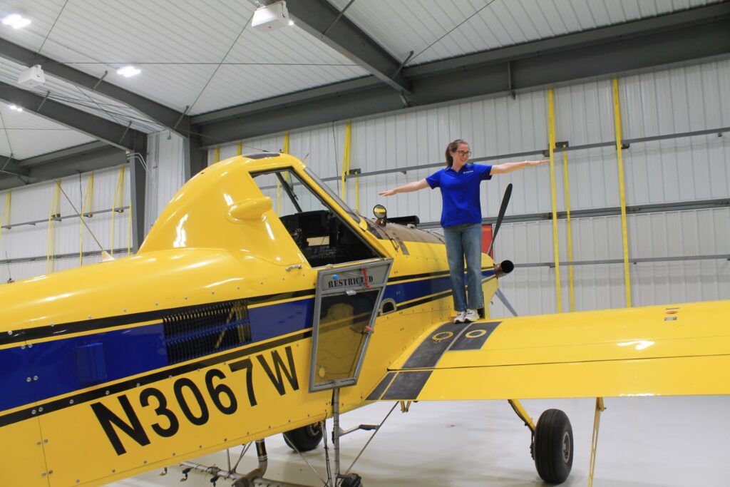 A woman stands on the wing of a yellow airplane with her arms extended on both sides.